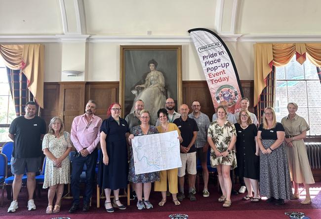 Group photograph of the Pride in Place Wick and Toddington Neighbourhood Board and project team standing together in the Littlehampton Town Council's Chamber, holding a map of the proposed Pride in Place neighbourhood boundary beside a Pride in Place event banner